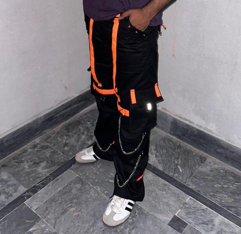 May include: Black cargo pants with orange accents, featuring multiple pockets and silver chain detailing. The pants have a reflective patch on the cargo pocket and are paired with white and grey sneakers.