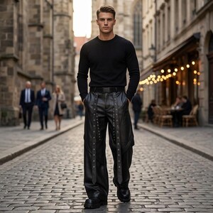 Men's wide-leg black leather motorcycle pants with decorative eyelet straps and industrial style, styled with a black sweater for an urban techwear look.