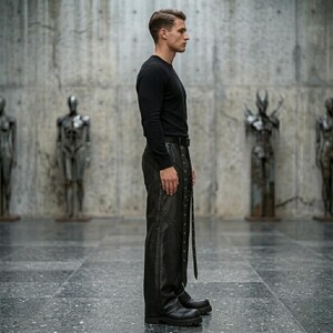 Side profile of baggy black leather pants showing the relaxed fit, premium leather texture, and the flow of the decorative grommet straps from waist to hem.