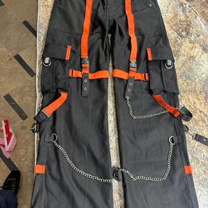 May include: Black cargo pants with orange straps and accents. The pants feature multiple pockets, metal chain details, and adjustable straps. The design includes silver metal hardware and a button closure.