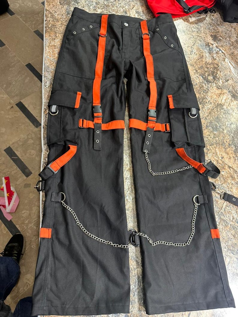 May include: Black cargo pants with orange straps and accents. The pants feature multiple pockets, metal chain details, and adjustable straps. The design includes silver metal hardware and a button closure.
