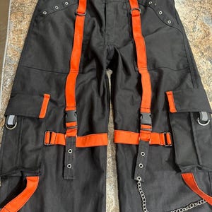 May include: Black cargo pants with orange straps and accents. The pants feature multiple pockets, metal hardware, and a silver chain detail. The orange straps have black plastic buckles. The pants are laid flat.