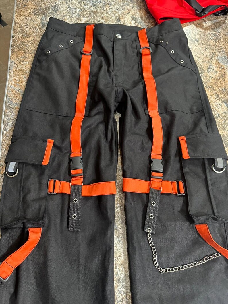 May include: Black cargo pants with orange straps and accents. The pants feature multiple pockets, metal hardware, and a silver chain detail. The orange straps have black plastic buckles. The pants are laid flat.