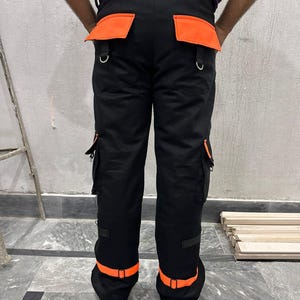 May include: Black work pants with orange accents. The pants feature orange pockets, belt loops, and ankle straps. The pants have multiple pockets and metal D-rings. The pants are made of a durable material.