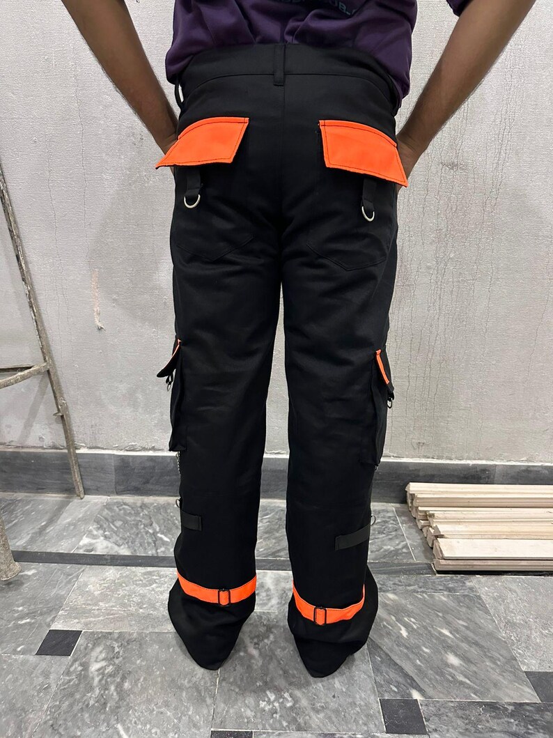 May include: Black work pants with orange accents. The pants feature orange pockets, belt loops, and ankle straps. The pants have multiple pockets and metal D-rings. The pants are made of a durable material.
