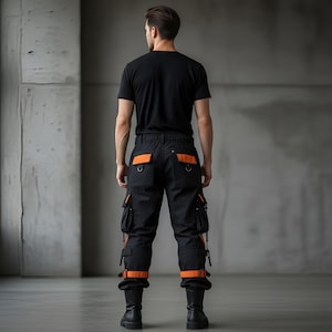 May include: Black cargo pants with orange accents, featuring multiple pockets and straps. The pants have orange detailing on the back pockets and around the ankles. The person is wearing a black t-shirt and black boots.