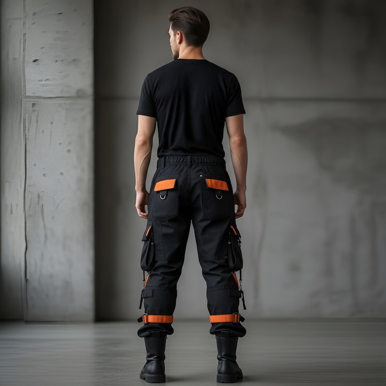 May include: Black cargo pants with orange accents, featuring multiple pockets and straps. The pants have orange detailing on the back pockets and around the ankles. The person is wearing a black t-shirt and black boots.