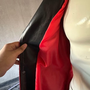 Pu&ograve; includere: Giacca in pelle nera con fodera rosso vivo. La giacca &egrave; aperta, rivelando il contrasto tra l'elegante esterno nero e l'interno rosso brillante. Una mano tiene aperta la giacca, mostrando il design.
