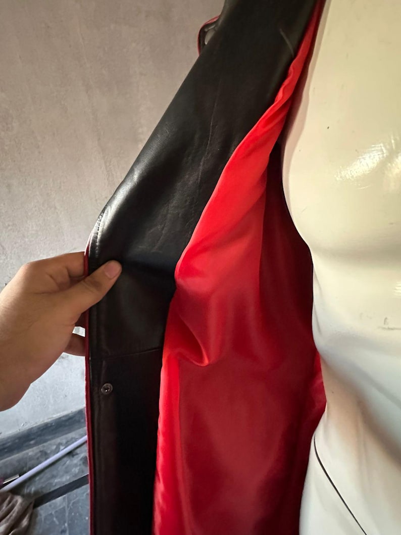 Pu&ograve; includere: Giacca in pelle nera con fodera rosso vivo. La giacca &egrave; aperta, rivelando il contrasto tra l'elegante esterno nero e l'interno rosso brillante. Una mano tiene aperta la giacca, mostrando il design.