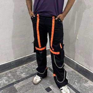 May include: Black cargo pants with bright orange straps and accents. The pants feature multiple pockets, silver chain details, and are paired with white and gray sneakers. A modern, urban style.