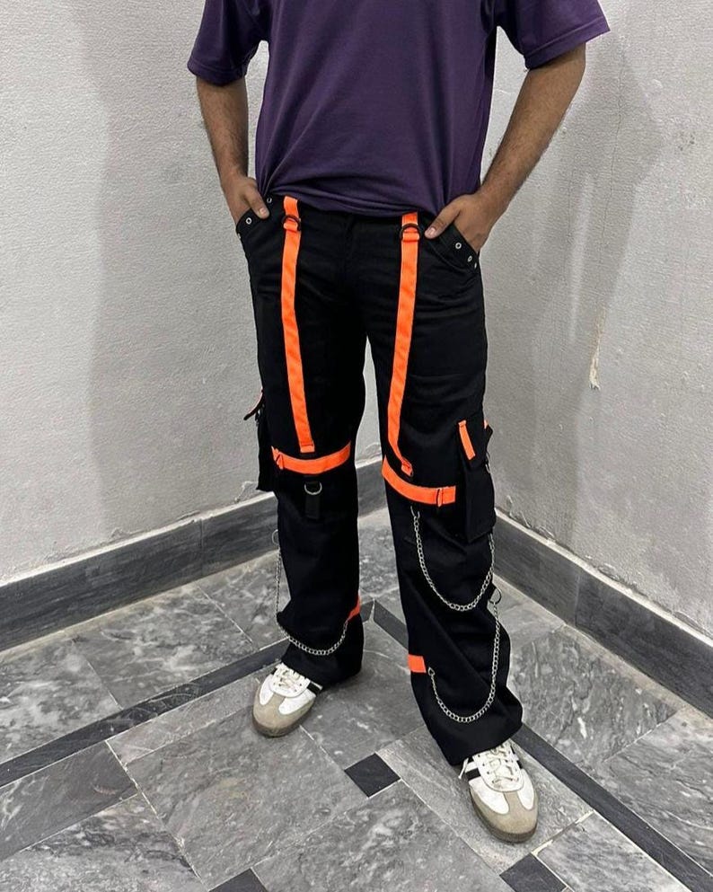 May include: Black cargo pants with bright orange straps and accents. The pants feature multiple pockets, silver chain details, and are paired with white and gray sneakers. A modern, urban style.