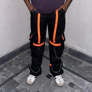 May include: Black cargo pants with bright orange stripes and accents. The pants feature multiple pockets, silver chain details, and are worn with white sneakers. A modern, urban style.