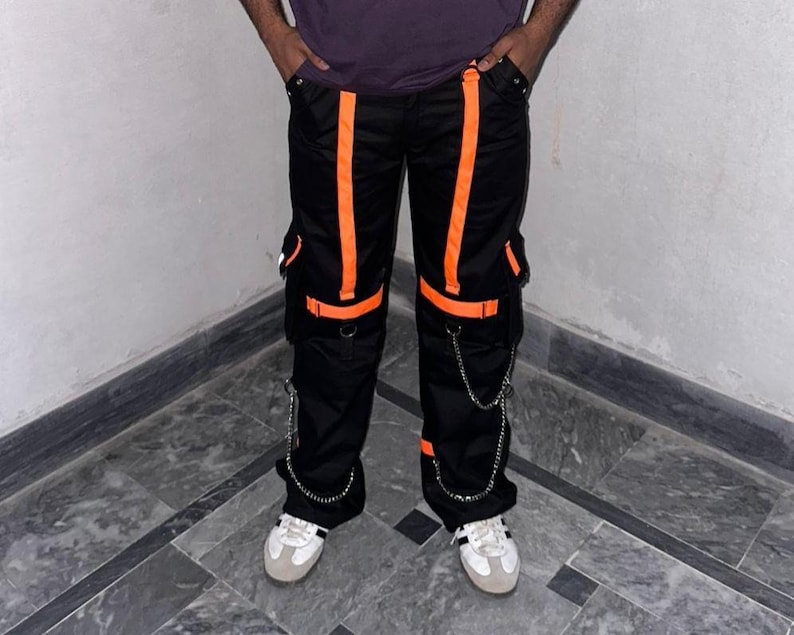 May include: Black cargo pants with bright orange stripes and accents. The pants feature multiple pockets, silver chain details, and are worn with white sneakers. A modern, urban style.