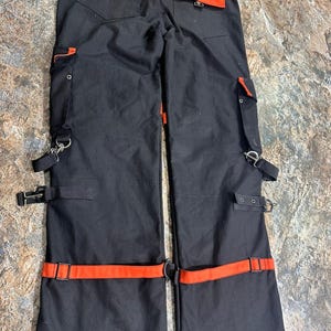 May include: Black pants with orange accents. The pants feature orange pockets, straps, and belt loops. The pants have multiple metal rings and buckles. The pants are made of a durable material.