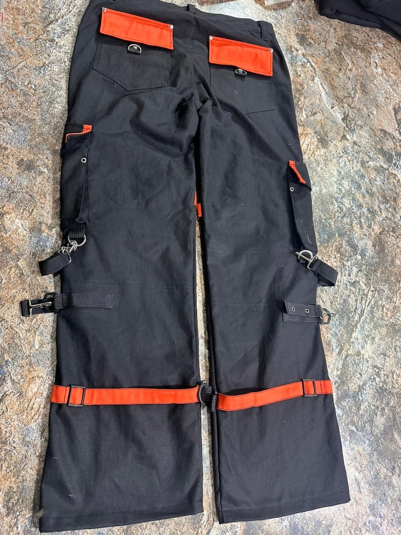 May include: Black pants with orange accents. The pants feature orange pockets, straps, and belt loops. The pants have multiple metal rings and buckles. The pants are made of a durable material.