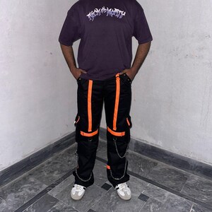 May include: A person wearing a dark purple t-shirt with the text "TRASHY" in a stylized font. The person is also wearing black cargo pants with orange stripes and accents, and white sneakers.