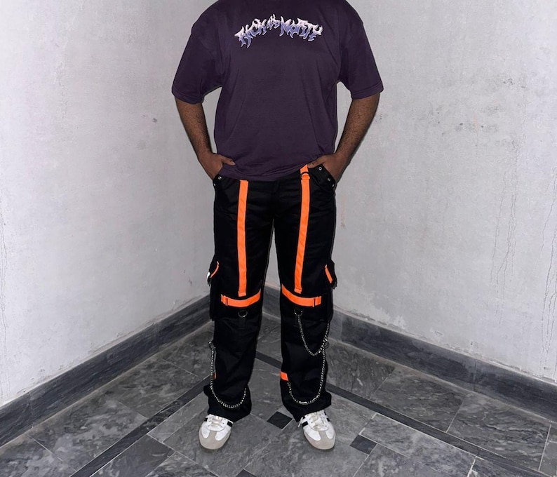 May include: A person wearing a dark purple t-shirt with the text "TRASHY" in a stylized font. The person is also wearing black cargo pants with orange stripes and accents, and white sneakers.