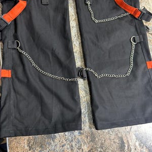 May include: Black cargo pants with orange accents, featuring multiple pockets, straps, and silver chain detailing. The pants have a modern, edgy design with a utilitarian aesthetic.