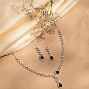 May include: A silver necklace and earring set with a teardrop design. The necklace and earrings feature dark blue gemstones surrounded by small, clear stones. The jewellery is displayed on cream fabric with a sprig of white flowers.