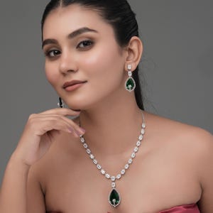 Puede incluir: Un deslumbrante conjunto de joyas con collar y pendientes. El collar tiene una serie de piedras claras de talla cuadrada que conducen a un colgante en forma de lágrima con una piedra central verde. Los pendientes reflejan el diseño del colgante.