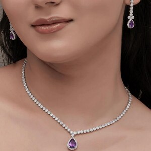 May include: A dazzling jewelry set featuring a necklace and earrings. The necklace and earrings have a silver-colored metal setting, adorned with numerous small, clear stones. Each piece showcases a teardrop-shaped, purple gemstone at its center, surrounded by a halo of smaller stones.