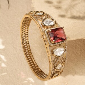 Peut inclure: Bracelet doré orné d'une grande pierre rouge carrée et de plusieurs petites pierres transparentes. Le bracelet présente un design ouvert avec des détails complexes et est serti de petits accents étincelants. Un bijou luxueux.