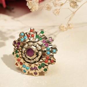 May include: A gold ring with a floral design, featuring a central purple gemstone and surrounded by small, colourful gems in red, green, blue, and purple. Tiny white pearls encircle the centre stone.