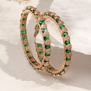 May include: Two gold bangles with emerald green rectangular stones and small, round, sparkling accents. The bangles are displayed on a white surface, with soft shadows and delicate dried flowers in the background.