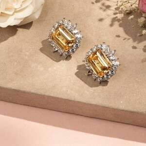 Puede incluir: Un par de pendientes de botón con una gema amarilla rectangular, rodeada de piedras claras más pequeñas. Los pendientes se muestran sobre una superficie neutra con flores. Joyas.