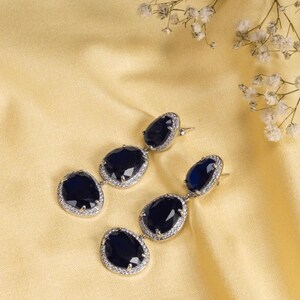 Puede incluir: Elegantes pendientes colgantes con tres piedras ovales de color azul oscuro, cada una rodeada por un halo de peque&ntilde;os cristales transparentes. Los pendientes se colocan sobre un fondo amarillo dorado suave, con delicadas flores blancas en la esquina superior derecha.