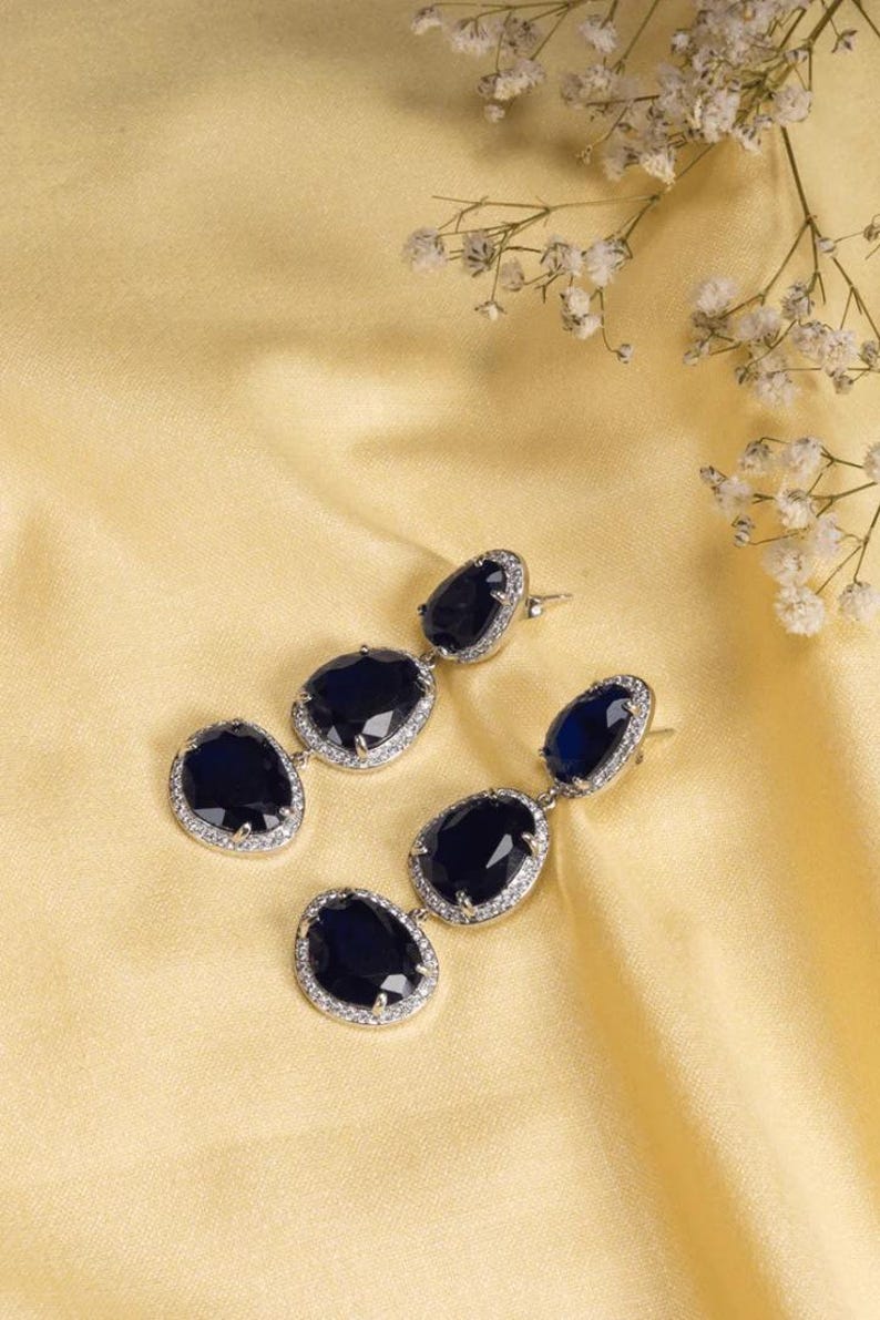 Puede incluir: Elegantes pendientes colgantes con tres piedras ovales de color azul oscuro, cada una rodeada por un halo de peque&ntilde;os cristales transparentes. Los pendientes se colocan sobre un fondo amarillo dorado suave, con delicadas flores blancas en la esquina superior derecha.