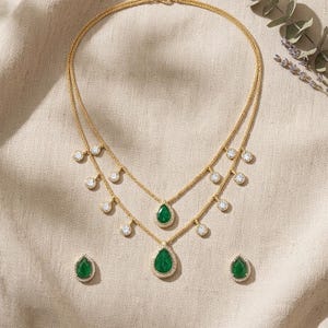May include: A gold-toned double-strand necklace featuring two teardrop-shaped green pendants and multiple small round clear stones along the chains, accompanied by a pair of matching teardrop-shaped green stud earrings with clear stone borders.