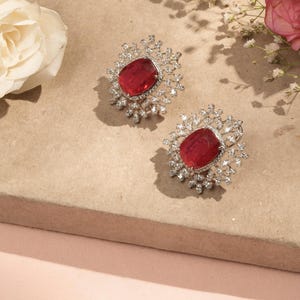 May include: A pair of silver-toned stud earrings. Each earring features a large, oval, ruby-red gemstone surrounded by a halo of smaller, clear crystals. The earrings are set against a neutral background with floral accents.