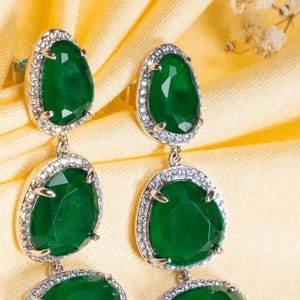 Puede incluir: Elegantes pendientes colgantes con tres piedras preciosas verde esmeralda facetadas, cada una rodeada por un halo de peque&ntilde;os cristales brillantes. Los pendientes cuelgan de un engaste plateado, creando un accesorio lujoso y llamativo.