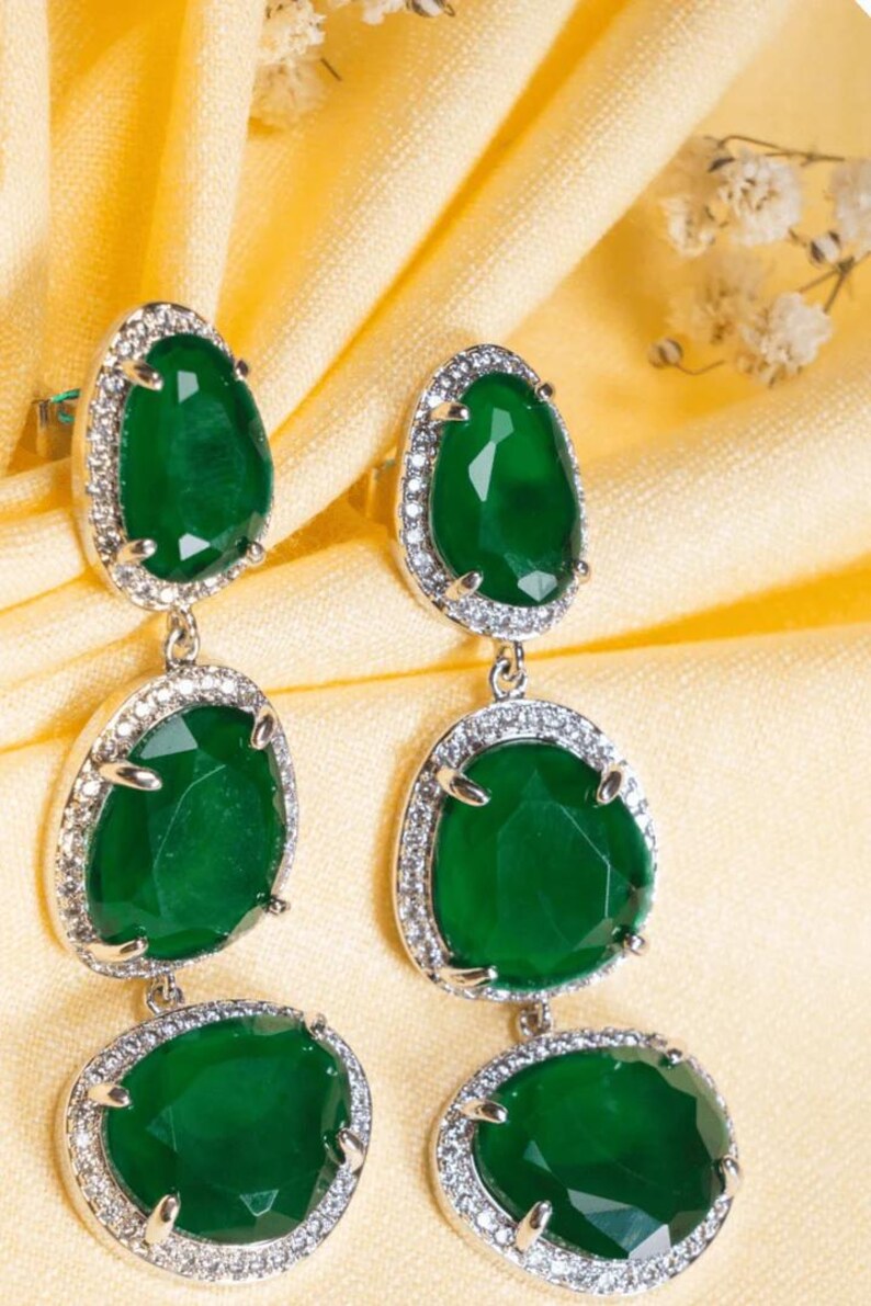Puede incluir: Elegantes pendientes colgantes con tres piedras preciosas verde esmeralda facetadas, cada una rodeada por un halo de peque&ntilde;os cristales brillantes. Los pendientes cuelgan de un engaste plateado, creando un accesorio lujoso y llamativo.