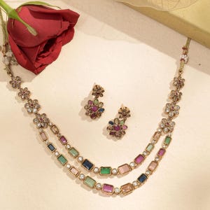 Puede incluir: Un conjunto de collar y pendientes dorados. El collar presenta dos hileras de piedras rectangulares de colores y detalles en forma de flor. Los pendientes tienen un diseño floral con piedras de colores. Una rosa roja está en el fondo.