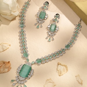 May include: A silver-toned necklace and matching earrings set. The jewelry features large, rectangular, light green gemstones surrounded by smaller clear stones. The necklace has a central pendant with a large gemstone and dangling accents.