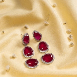 May include: A pair of ruby-red earrings with a silver-colored, diamond-studded border. The earrings are set against a yellow satin background, with small white flowers scattered around.