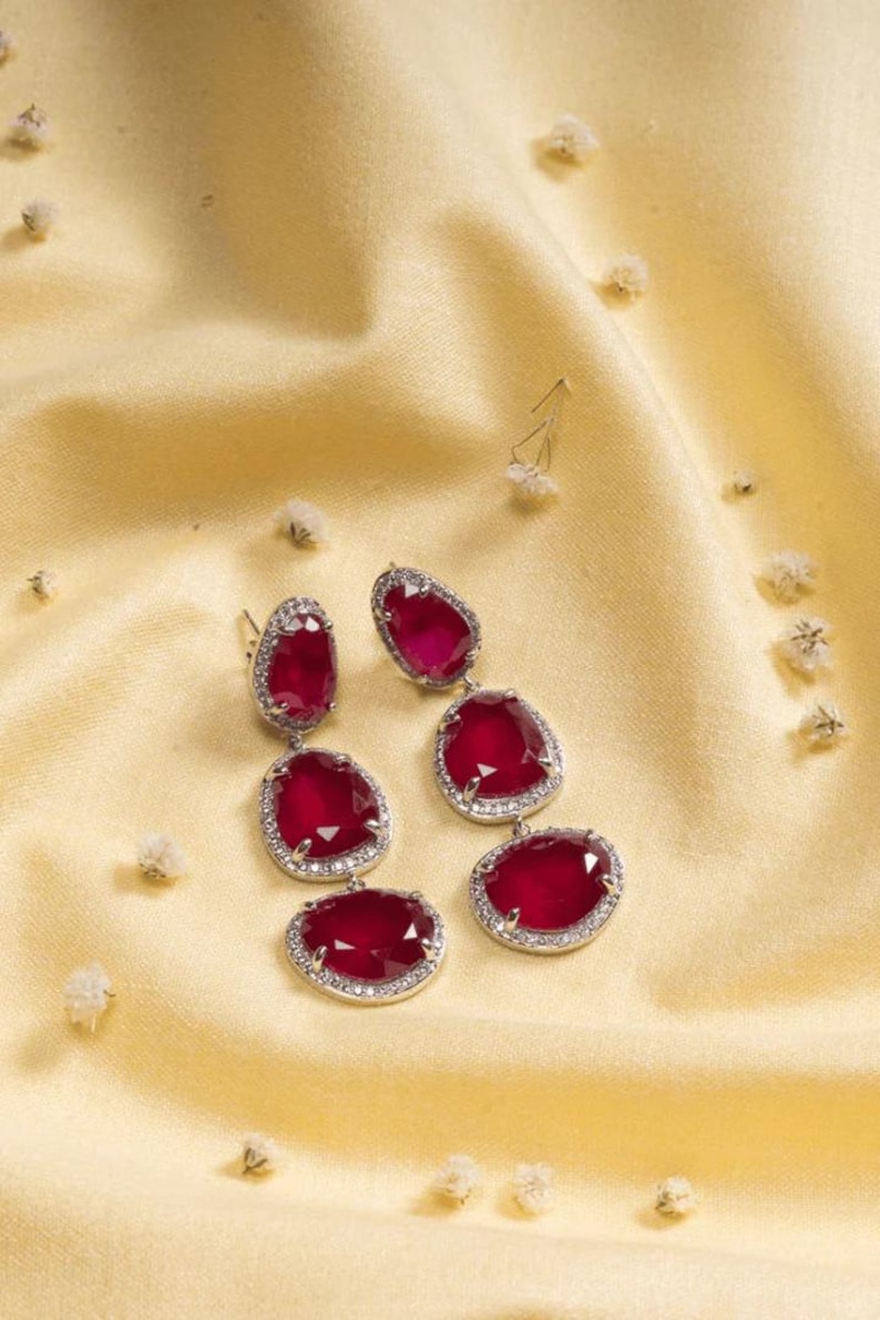 Puede incluir: Un par de pendientes de color rojo rub&iacute; con un borde plateado con incrustaciones de diamantes. Los pendientes est&aacute;n colocados sobre un fondo de sat&eacute;n amarillo, con peque&ntilde;as flores blancas esparcidas.