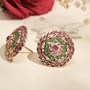 Può includere: Orecchini a lobo in oro rosa con un design circolare. Gli orecchini presentano un motivo di pietre preziose verdi, rosa e bianche. Gli orecchini sono posizionati su uno sfondo morbido e neutro.