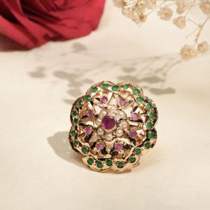 May include: A gold-toned ring with a floral design, featuring a central ruby surrounded by small pearls and alternating green and pink gemstones. The ring is set against a soft, off-white background with a red rose and delicate white flowers.