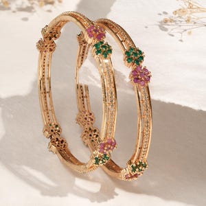 Può includere: Due braccialetti color oro con decorazioni floreali. Ogni braccialetto presenta piccole pietre preziose rosa e verdi ed è adornato con minuscole pietre trasparenti. I braccialetti sono esposti su una superficie bianca.