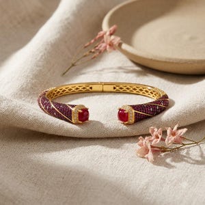 May include: A gold cuff bracelet featuring a hinged design with a lattice pattern on the inner band. The exterior is encrusted with deep red pavé stones and diagonal rows of clear crystals, finished with a large, round, cabochon-cut red stone.