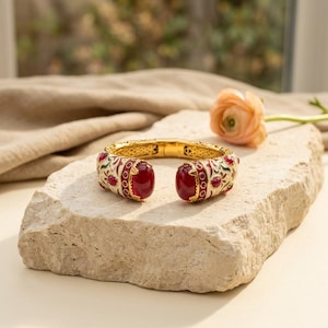 May include: A gold-toned hinged cuff bracelet featuring a white enamel base with intricate floral patterns in green and red. The bracelet is accented with large, smooth, deep red cabochon stones at the opening and smaller red stones along the band.