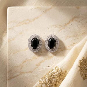 May include: A pair of oval-shaped stud earrings featuring a central black gemstone surrounded by a halo of small, clear, sparkling stones set in a silver-toned metal frame. The earrings have a classic design with a polished finish and secure backings.