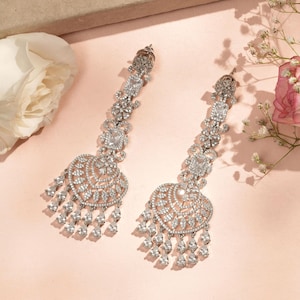 May include: A pair of silver drop earrings. Each earring has a long, decorative design with multiple clear, sparkling stones. The bottom of each earring features a heart-shaped design with dangling teardrop-shaped stones. The earrings are set against a soft pink background.