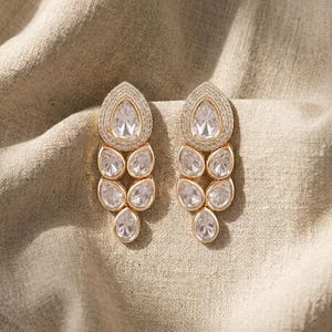 May include: A pair of gold-toned drop earrings featuring a cluster of clear, pear-shaped stones. Each earring has a large top stone surrounded by a halo of small pavé crystals, followed by four smaller pear-shaped stones arranged in a vertical drop.