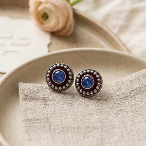 May include: A pair of round stud earrings featuring a central faceted blue stone surrounded by a deep red enamel ring and an outer border of silver-toned metal beads. The design includes a twisted rope texture detail between the enamel and the beads.
