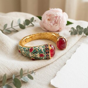 May include: A gold-tone hinged cuff bracelet featuring a teal and white enamel floral pattern, accented with multiple smooth, round, deep red cabochon stones. The bracelet has a wide, open-ended design with a secure clasp mechanism.