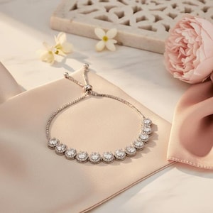 May include: Silver-toned bracelet with a row of round, clear gemstones. The bracelet has an adjustable sliding clasp and is displayed on a light pink satin surface, with flowers in the background.